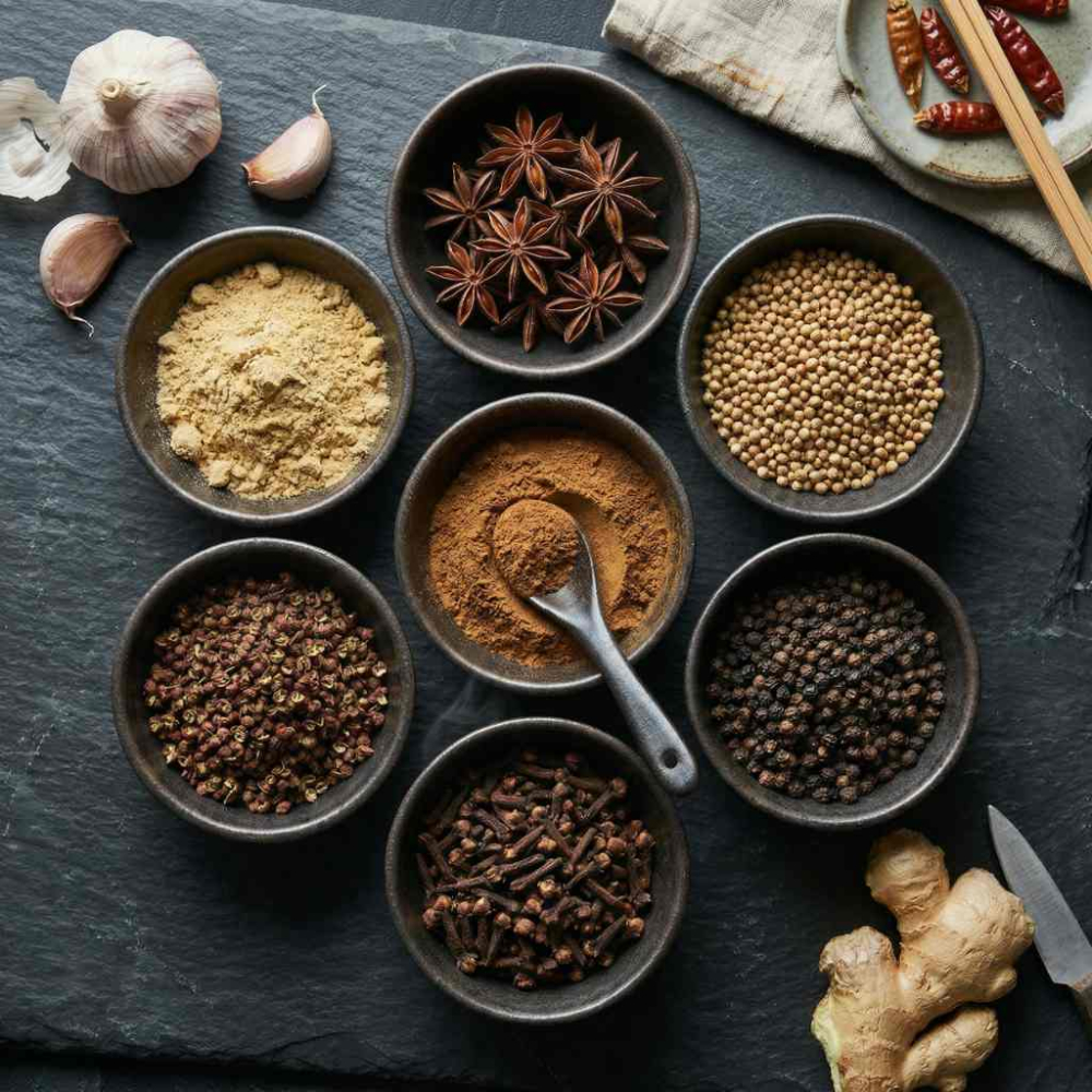 flat lay of Chinese spices on a dark stone surface, small ceramic bowls filled with ground ginger, star anise, coriander seeds, black peppercorns, cloves, Sichuan pepper, and five spice powder, fresh garlic and ginger root placed casually