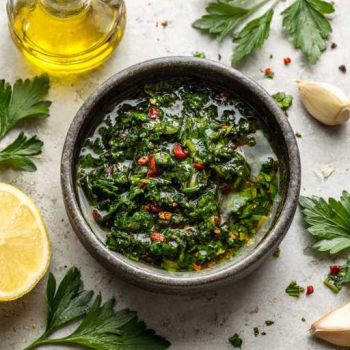 resh homemade chimichurri in a small matte ceramic bowl on a cool pale stone surface. The chimichurri is vibrant green with visible chopped flat-leaf parsley, fresh oregano, minced garlic, red chilli flakes, and glossy olive oil pooling slightly on top. A