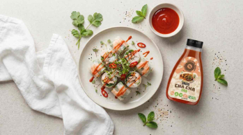 fresh Vietnamese style rice paper spring rolls on a pale ceramic plate, cut into bite sized pieces, topped with microgreens and thin slices of red chili and light drizzle of sriracha sauce, small ceramic dipping bowl filled with bright red sriracha sauce,